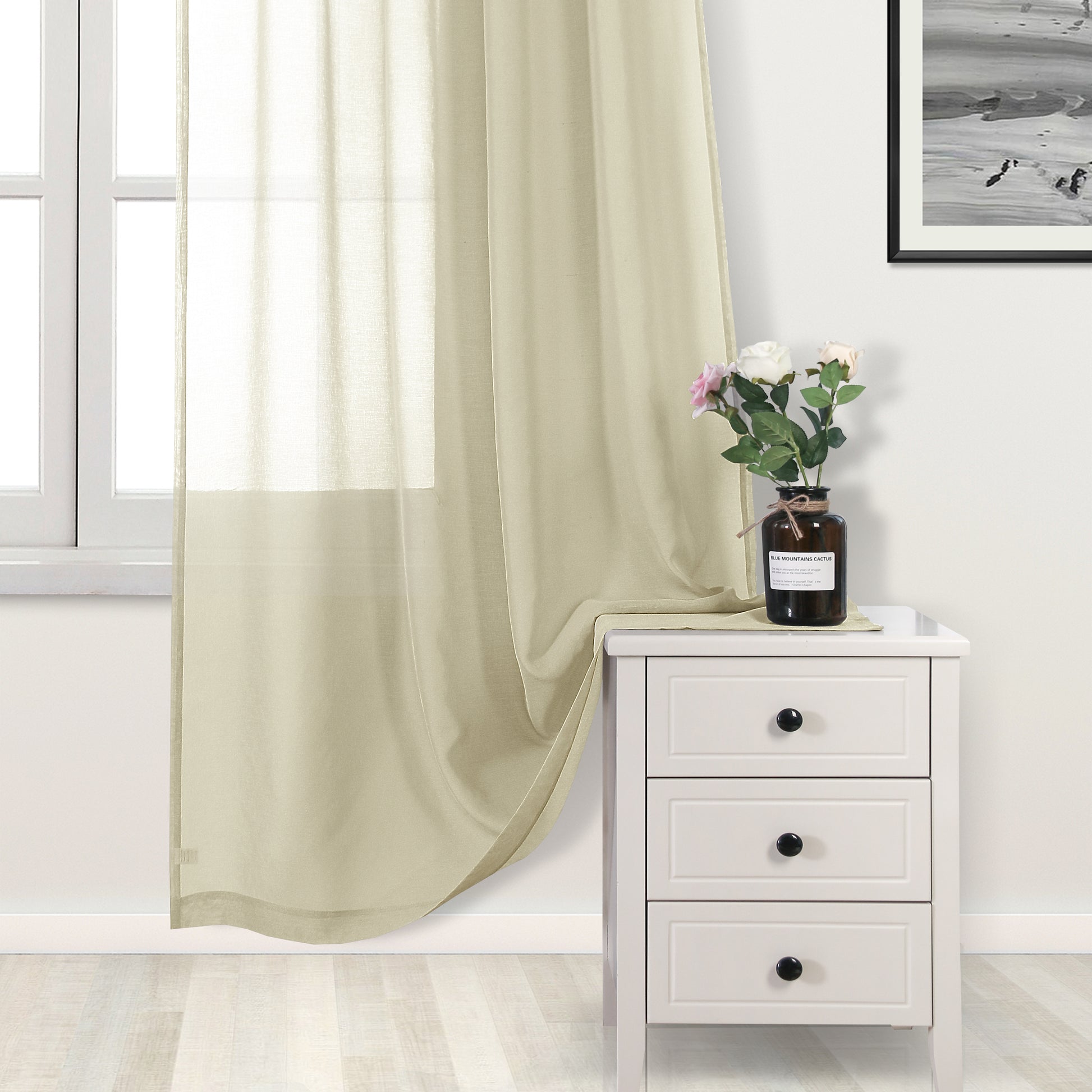 Beige curtain partially drawn over a window with a white nightstand and vase of flowers in the foreground.