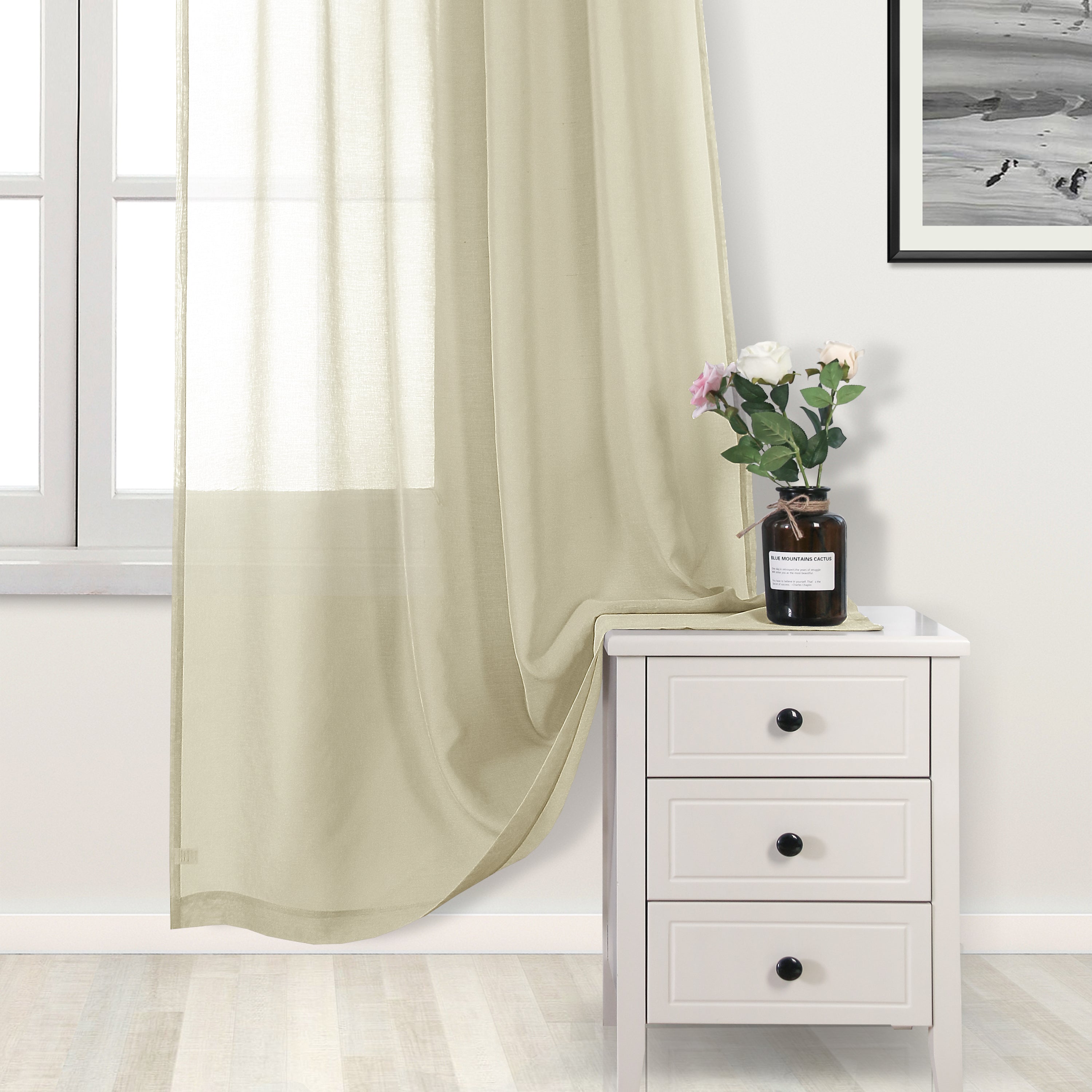 Beige curtain partially drawn over a window with a white nightstand and vase of flowers in the foreground.